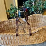 Buff Moses basket with brown leather handles on a carpeted floor in front of a fire place.