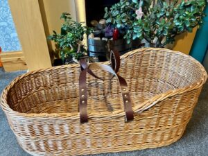 Buff Moses basket with brown leather handles on a carpeted floor in front of a fire place.