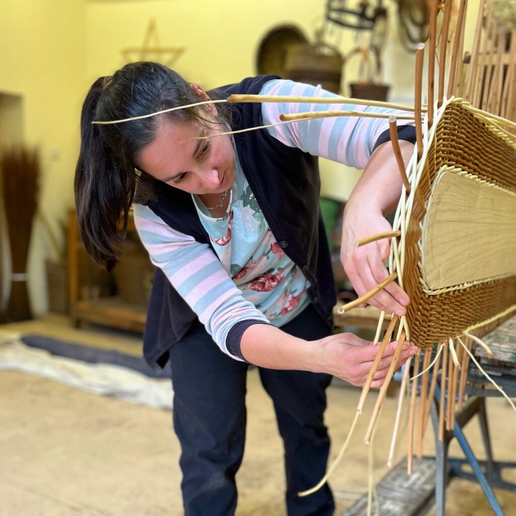 Steph wearing a flowery top, leaning over to weave around the central piece of wood on a coffin lid, using white willow to weave.