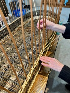 Action photo of lady helping to weave her own coffin moving a white willow rod behind an upright