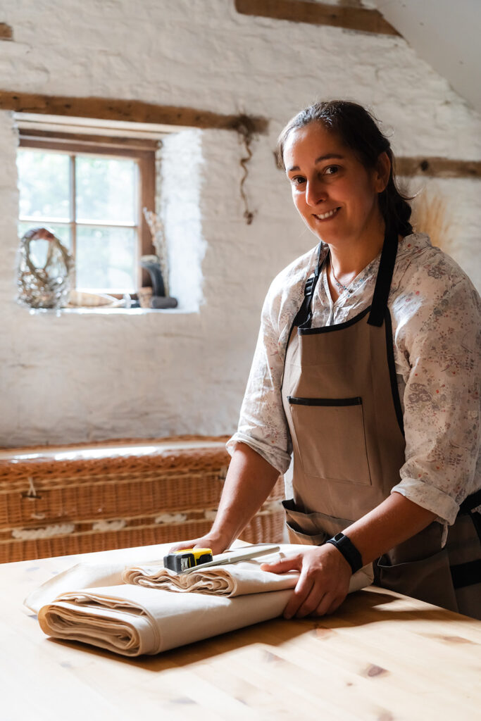 Steph in workshop with willow coffin behind and holding calico and sewing materials in hands on table