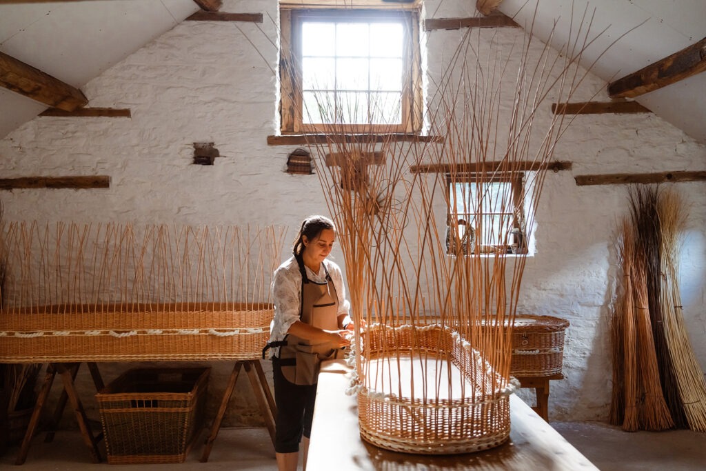 Steph working on a willow coffin in the rustic workshop with 2 coffins behind her, and sunlight coming in from the top window. 